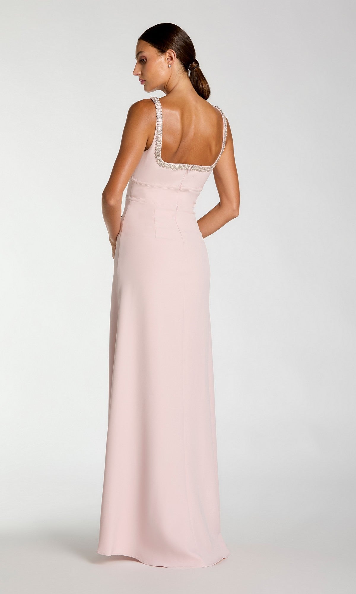 A woman stands with her back to the camera against a plain backdrop, wearing the Long Formal Dress: Mac Duggal 27633—a sophisticated pale pink crepe gown with a square neckline and back, elegantly finished with rhinestone trim. This refined silhouette captures the timeless glamour inherent in Mac Duggal designs.
